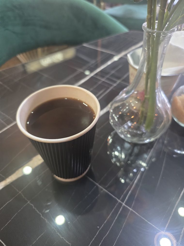 A ribbed takeaway-style cup of black coffee beside a small bud vase